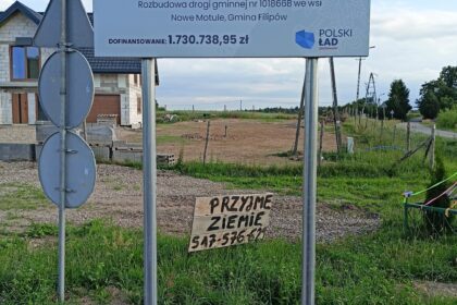 Tablica o rozbudowie drogi w gminie Filipów z funduszu Polski Ład. Pod nią odręczny napis na drewnie: Przyjmę ziemię