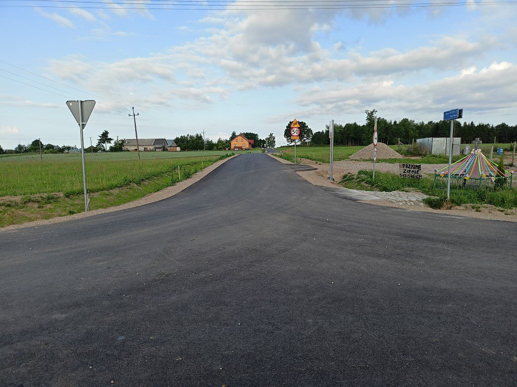 Nowa droga asfaltowa w wiejskiej okolicy z widocznymi znakami drogowymi i przydrożną kapliczką