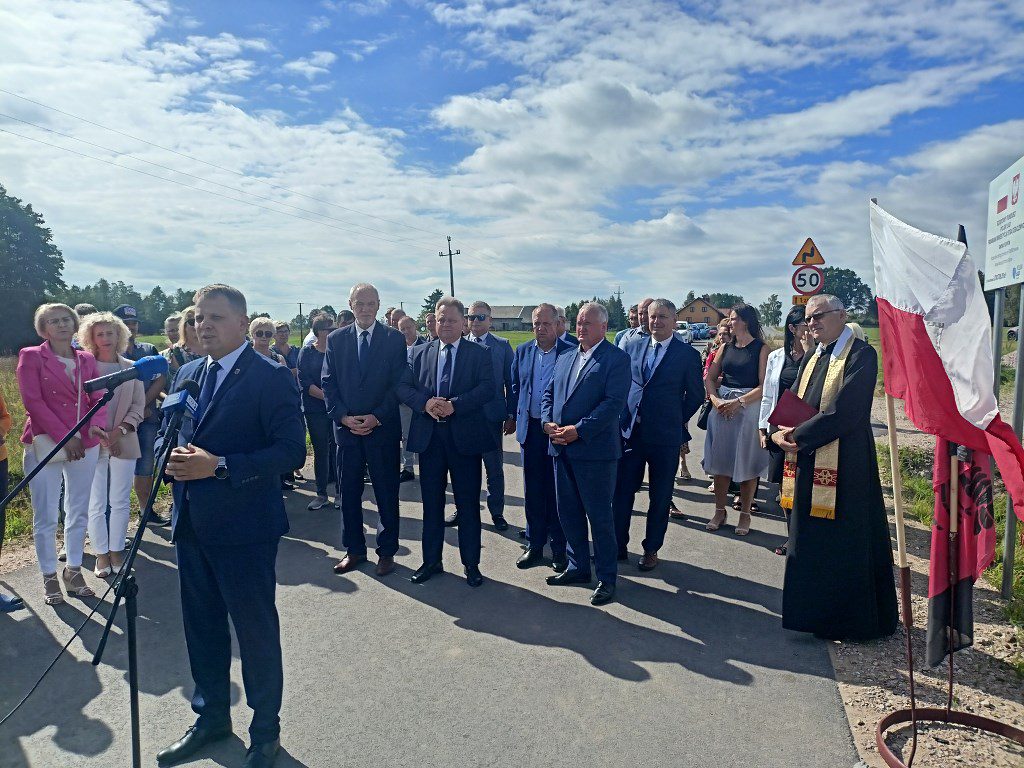 Mężczyzna przemawia do mikrofonu podczas uroczystego otwarcia drogi w obecności urzędników i księdza
