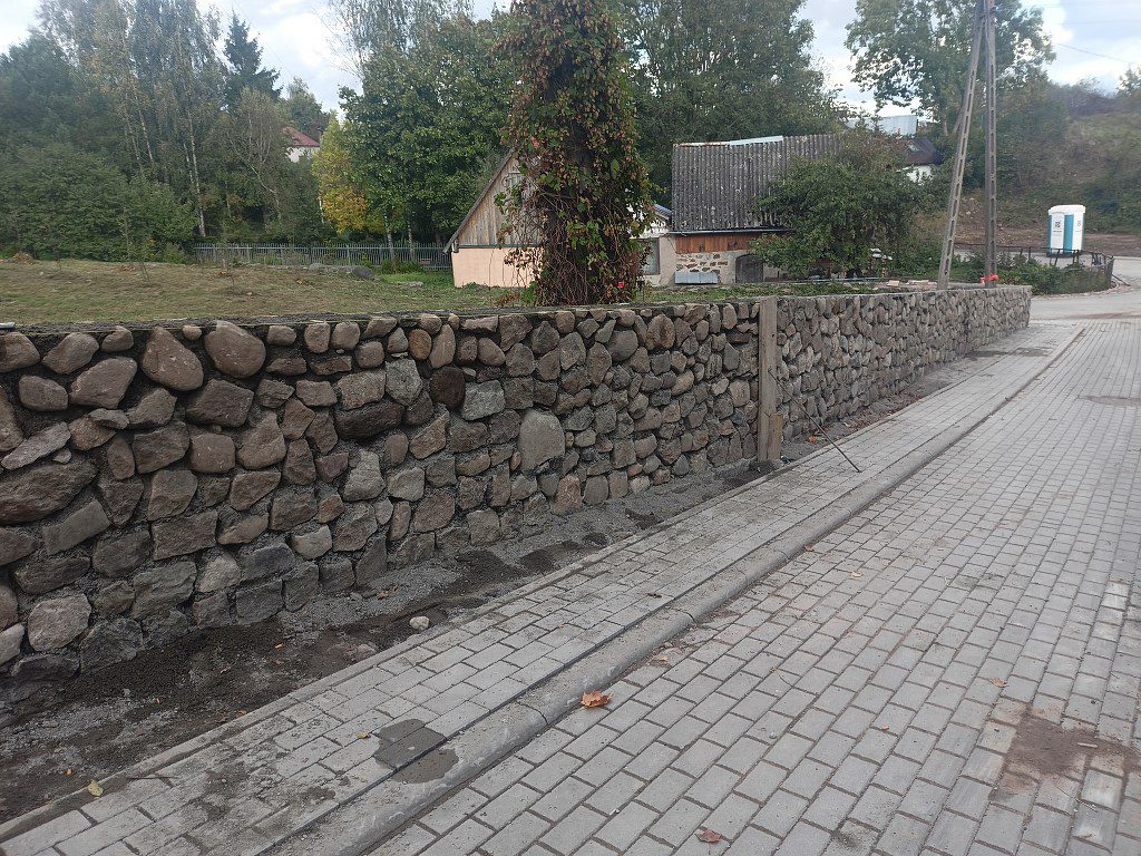 Mur z kamienia polnego wzdłuż chodnika z kostki brukowej na tle wiejskich zabudowań i drzew